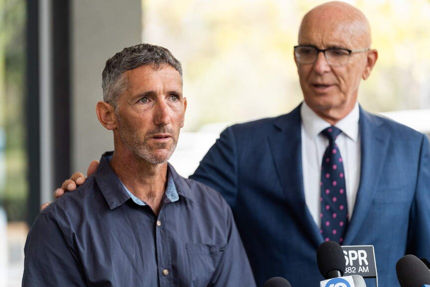 Aaron Cockman speaking to the media with John Quigley standing next to him, with Mr Quigley's hand on his shoulder.