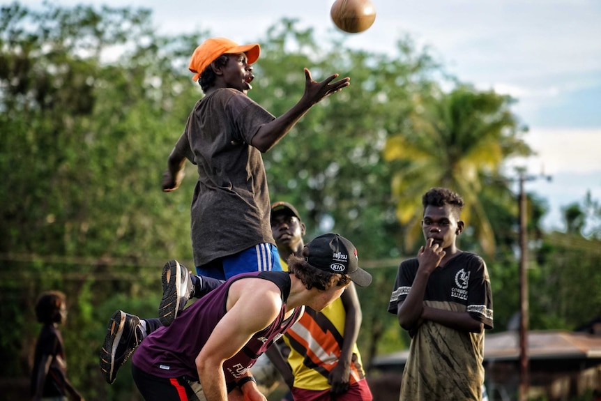 A young Tiwi footballer rides the back of an AFL player to catch the ball.
