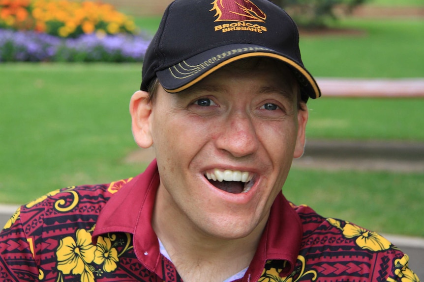 A man wearing a Brisbane Broncos cap and shirt sits smiling in a park.