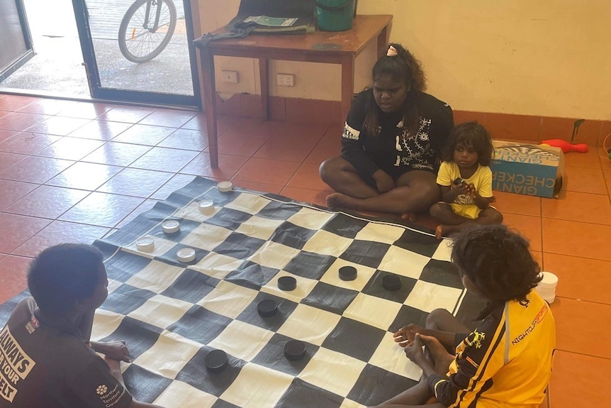 an aboriginal woman playing chess with three kids on a floor
