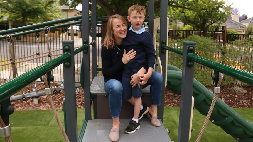 A picture of a woman sitting on school play equipment smiling, while she holds a boy in a blue school uniforms, who stands .