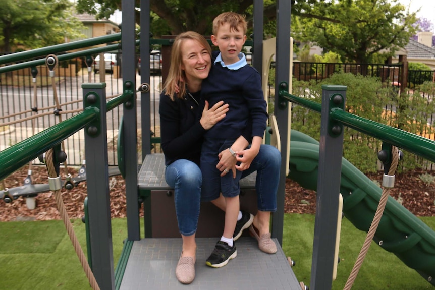 A picture of a woman sitting on school play equipment smiling, while she holds a boy in a blue school uniforms, who stands .