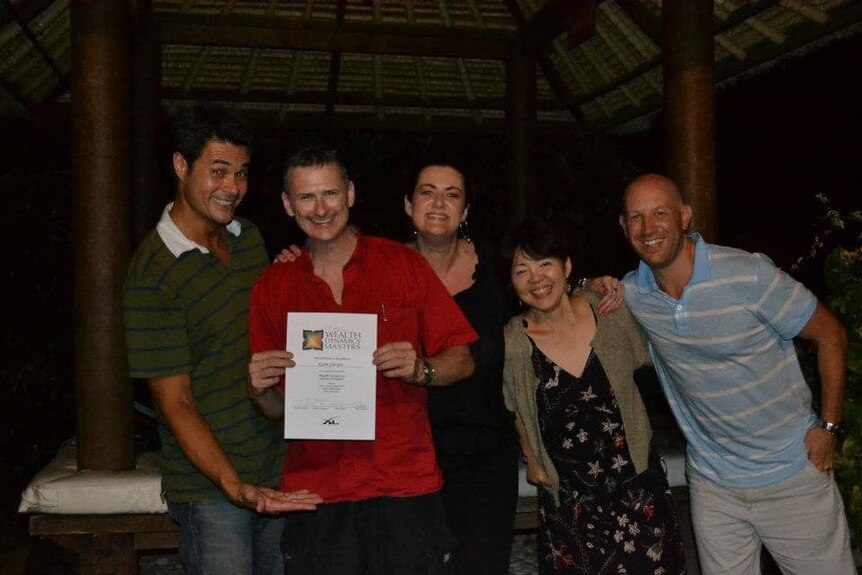 A photo of Karm Gilelspie wearing a red shirt standing  with several friends holding his graduation certificate.