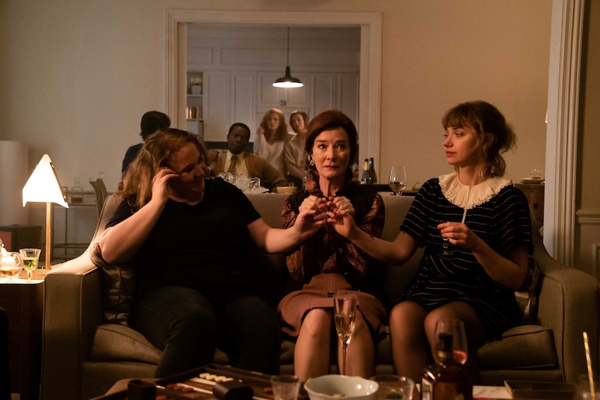 A scene from the film French Exit with Danielle Macdonald, Valerie Mahaffey and Imogen Poots sitting on a couch in an apartment