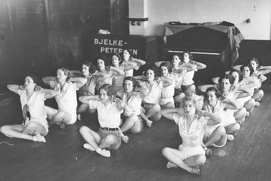 A black and white image show three lines of women sit cross-legged on the floor with arms out and elbows up.
