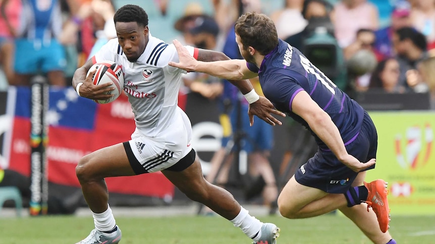 A man runs with a rugby ball under his right arm whilst another man runs towards him with his arm outstretched.