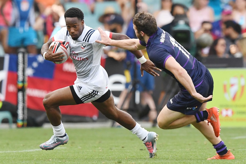 A man runs with a rugby ball under his right arm whilst another man runs towards him with his arm outstretched.