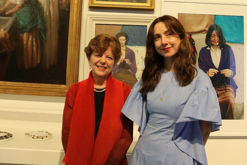 Two women pose in front of a wall decorated with art.