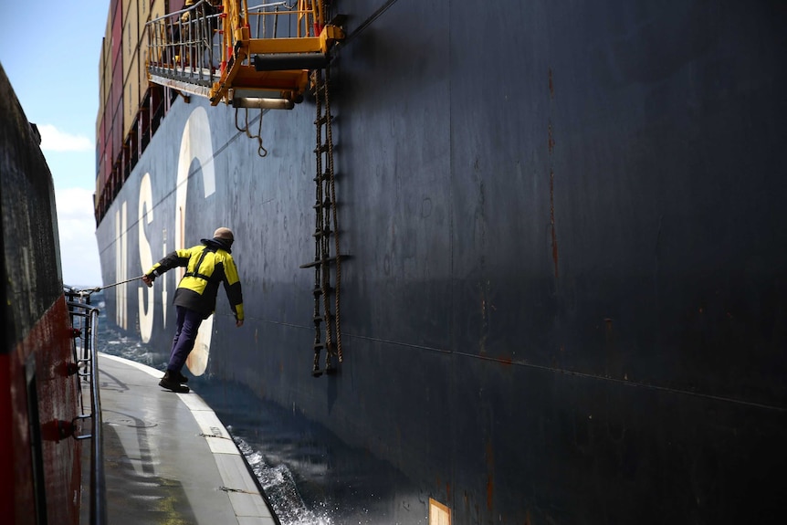 A man stands on a small boat next to a tanker ship with a rope ladder on the side.