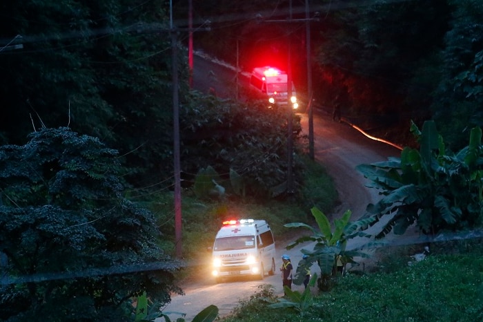 View from a distance at night as two ambulances with lights flashing drive down a winding unsealed road surrounded by lush trees