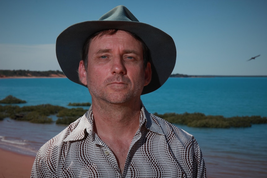 A man in a wide brim hat standing in front of a beach background