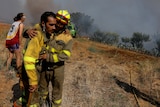 A firefighter in a yellow suit leans on another as smoke fills the air behind them. 