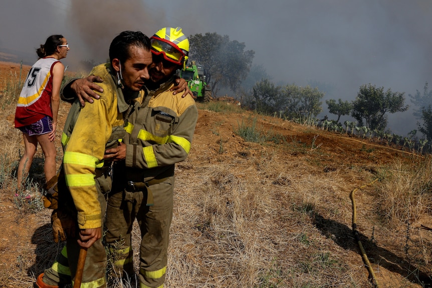 A firefighter in a yellow suit leans on another as smoke fills the air behind them. 