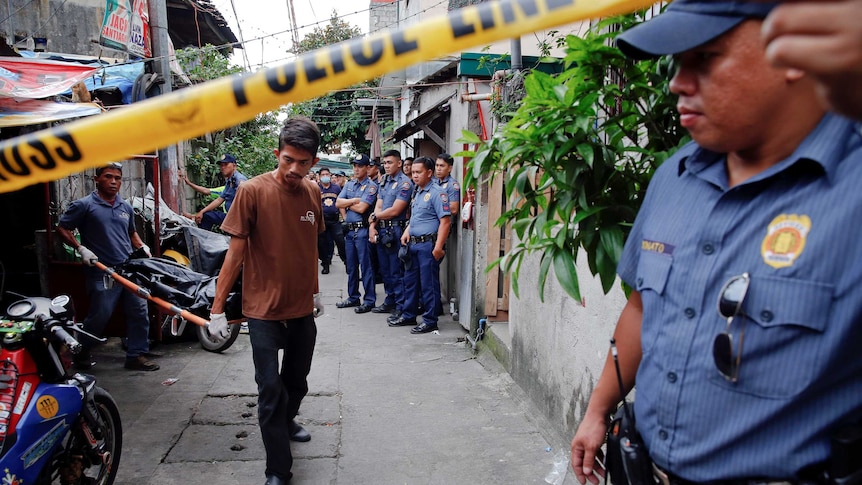 Policemen stand in an alley next to yellow and black police tape, as two bodies are wheeled out.