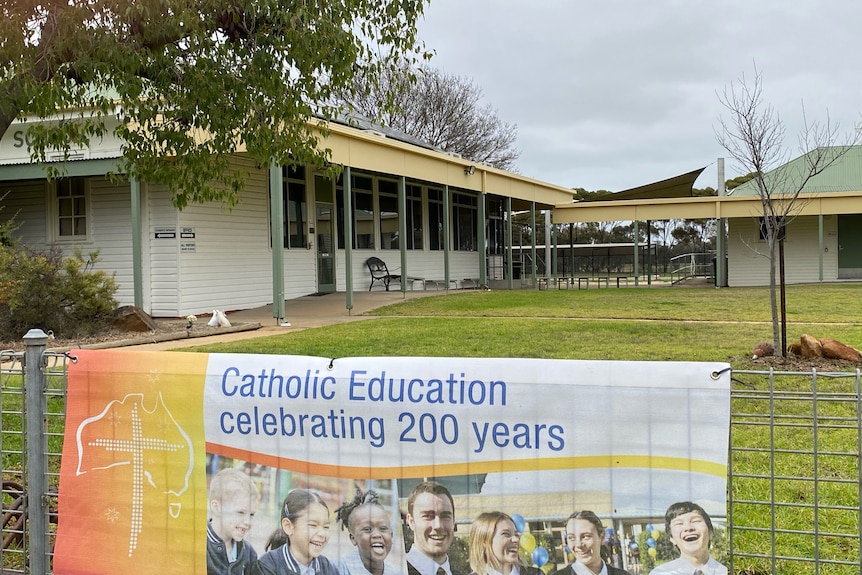 The view of a white wooden building. A sign on a fence reads "Catholic Education celebrating 200 yeras"