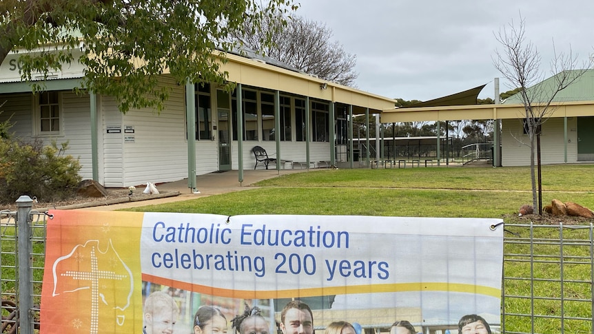 The view of a white wooden building. A sign on a fence reads "Catholic Education celebrating 200 yeras"