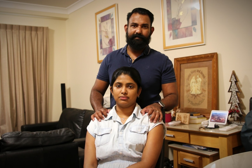 Neethu sits on a chair with her husband behind her, his hands on her shoulders.