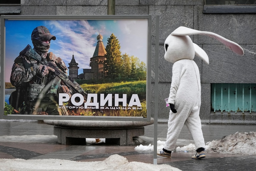 A street performer dressed up as a white rabbit walks past an army recruitment poster in St Petersburg