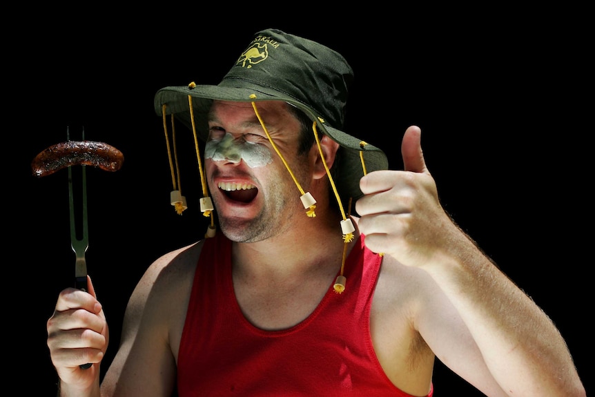 Man wearing a hat with corks wink while holding a barbecue fork with a sausage on it.