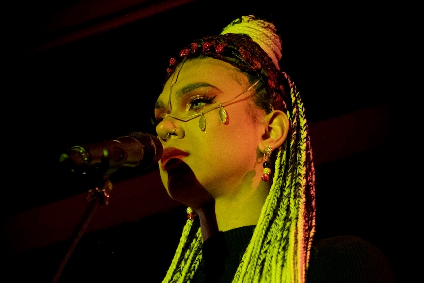 Jack is standing in front of a microphone. They are wearing long yellow braided hair and face jewellery.
