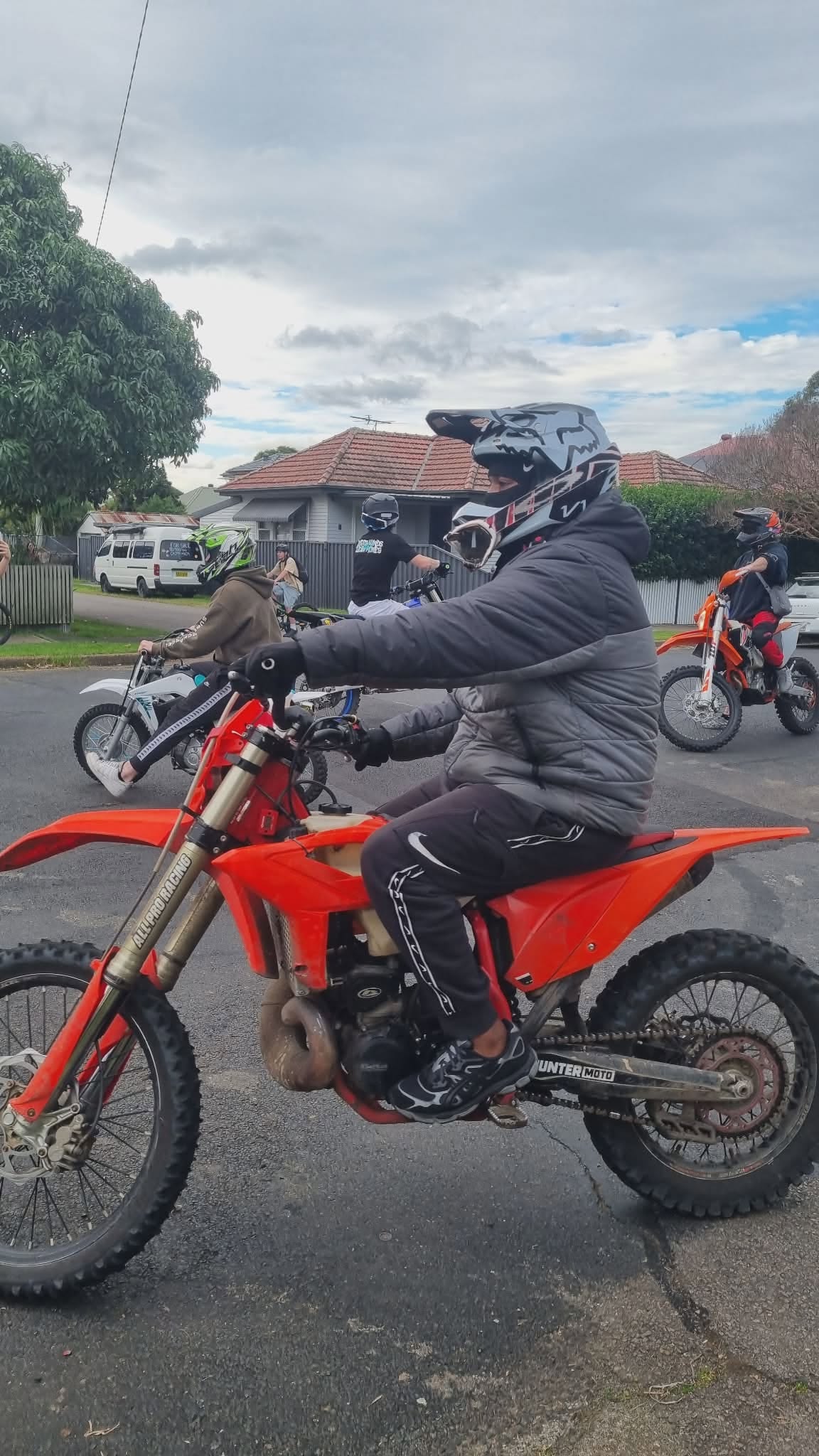Dirt bike riders wearing helmets riding around a suburban road with houses. 