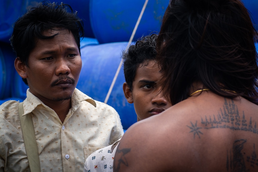 A man with shoulder length hair stands facing two men in button up shirts with serious expressions on fishing boat.