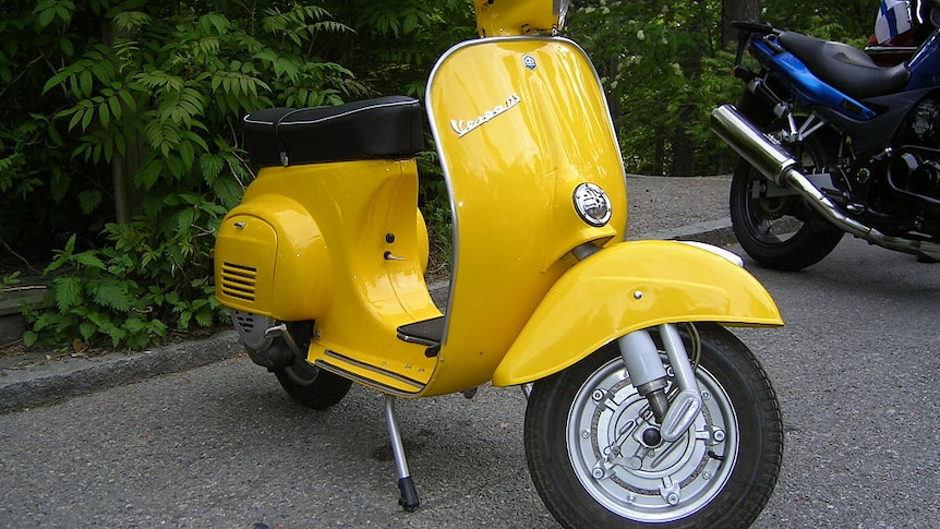 A bright yellow Vespa scooter parked in a parking lot in front of plants.