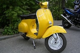 A bright yellow Vespa scooter parked in a parking lot in front of plants.