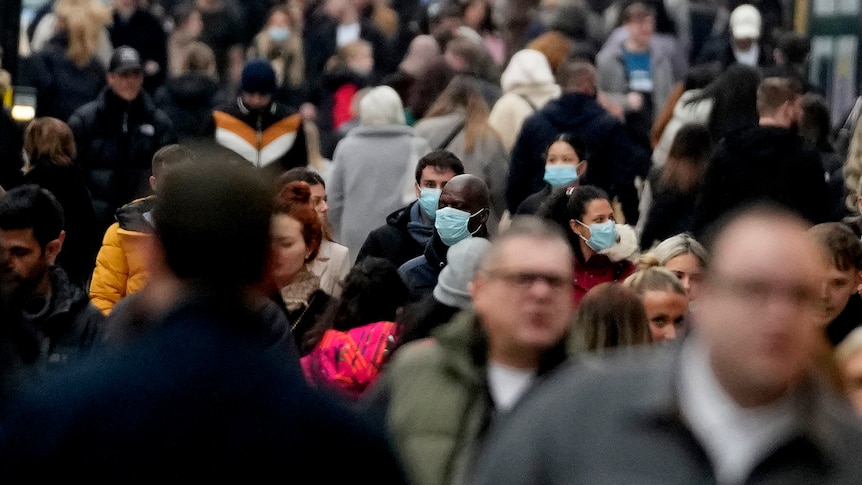 Dozens of people walk along a busy street in London, some wearing face masks