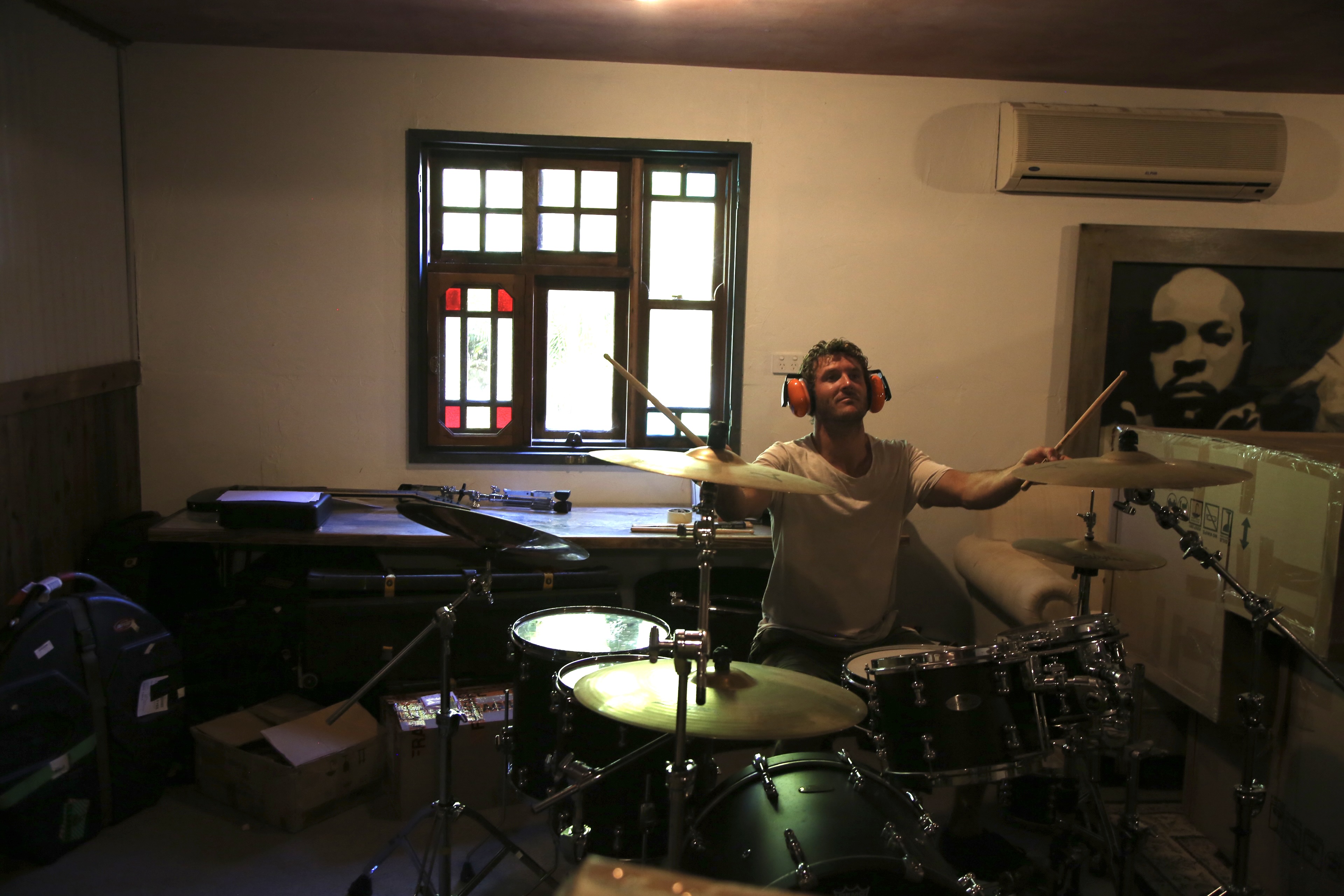 A man wears headphones while playing drums in a basement. A stainglass window is in background