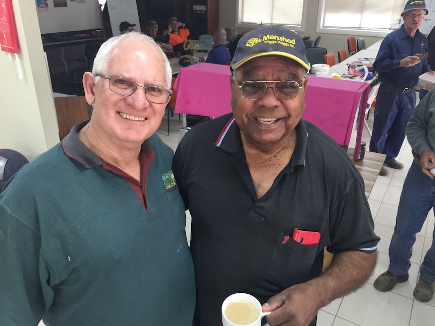 two men standing in the foreground, one with a cup of tea, with other men seated in the background