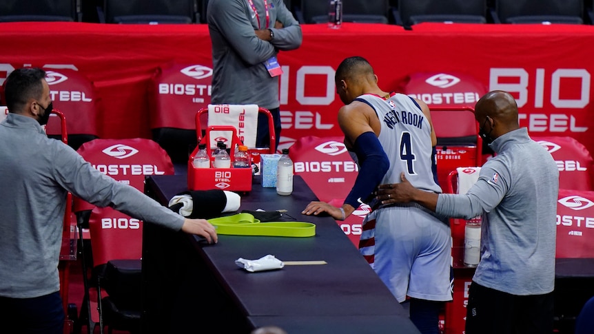 A man puts his arm around the waist of an injured NBA basketballer to help him out of the arena to the locker room. 