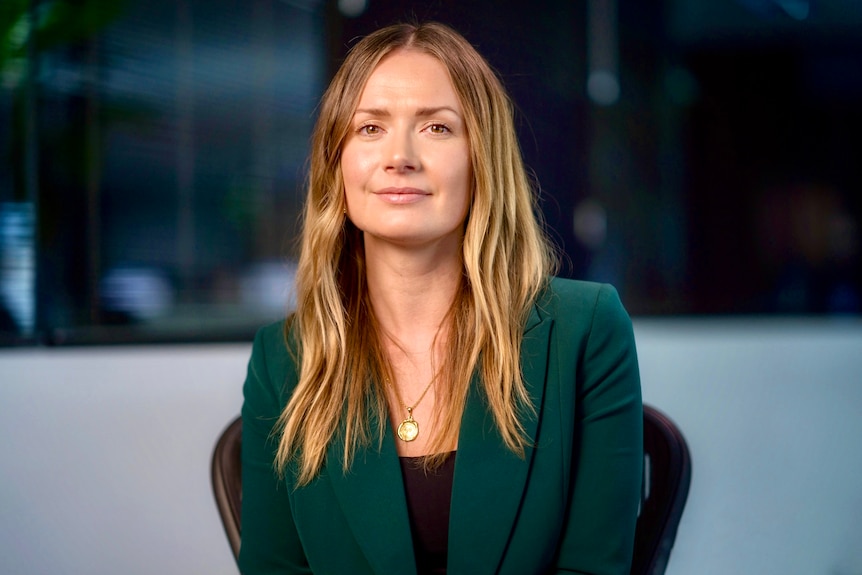 A woman with long hair sits on a chair in an office and looks into the camera.