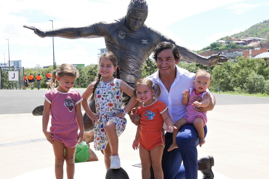 A man kneels with four young girls in front of a bronze statue of himself.