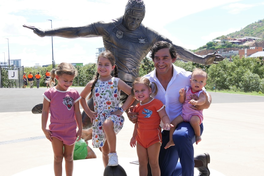 A man kneels with four young girls in front of a bronze statue of himself.