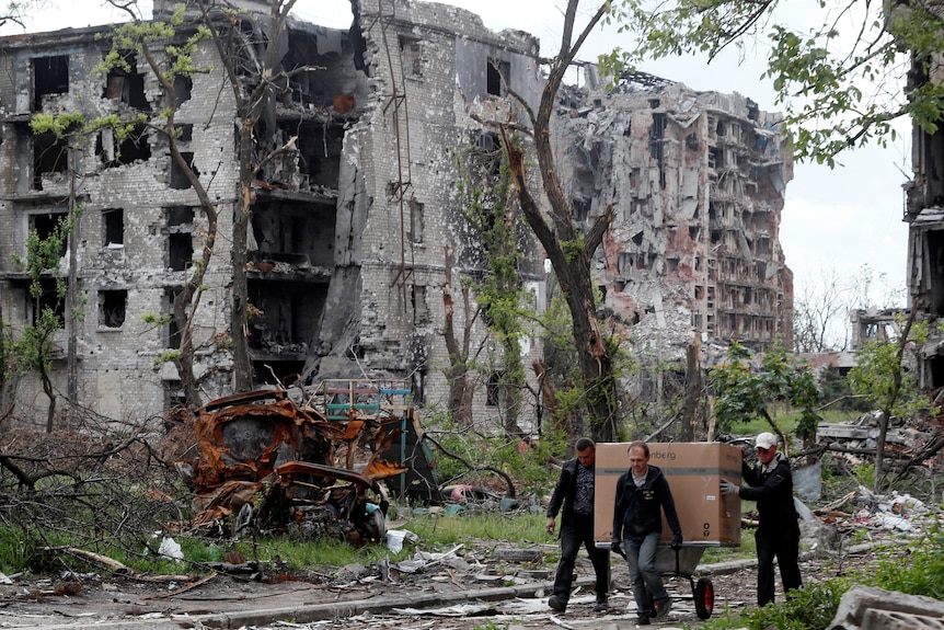 Local residents transport a box on a wheelbarrow past a heavily damaged apartment building near Azovstal Iron and Steel Works