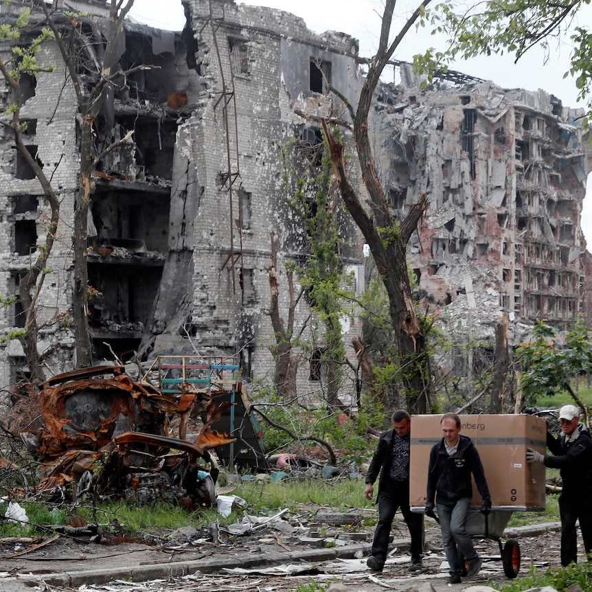 Local residents transport a box on a wheelbarrow past a heavily damaged apartment building near Azovstal Iron and Steel Works