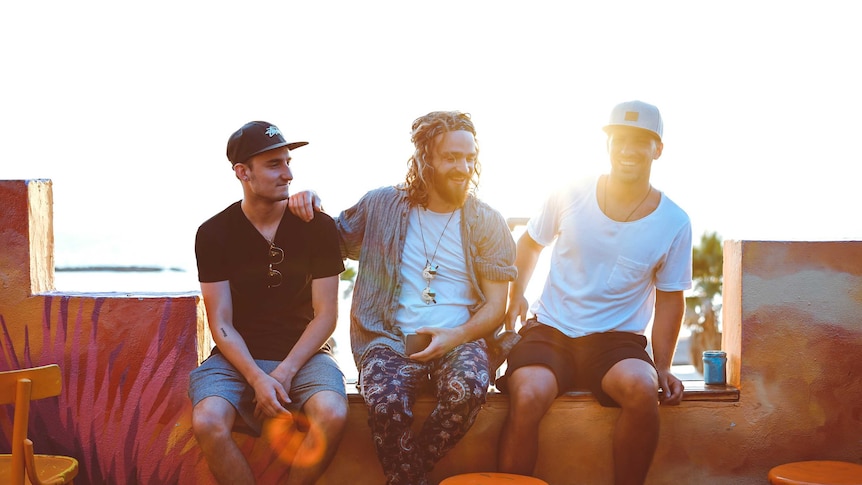 Three friends sitting next to each other outside on a ledge with the sun behind them to depict making friends as an adult