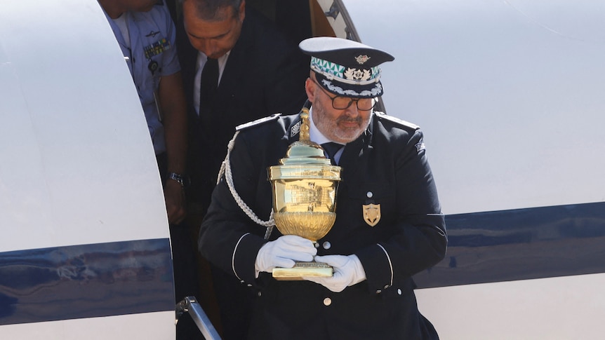 A man in a decorated, official-looking blue uniform carries a golden urn down the steps of an aeroplane.