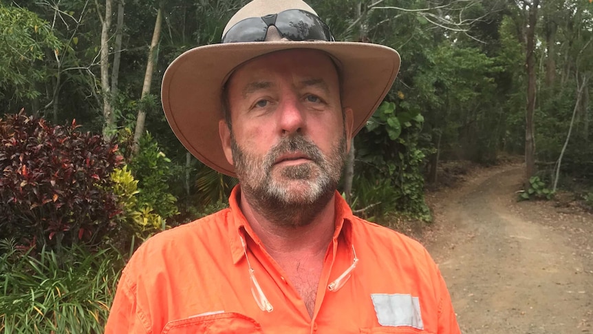 A man in a high-vis jacket, akubra and sunglasses looks tired as he stands on a dirt road in the bush.