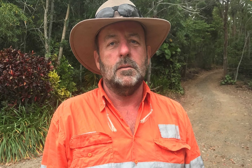 A man in a high-vis jacket, akubra and sunglasses looks tired as he stands on a dirt road in the bush.