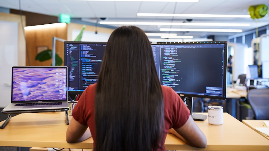 A person with long dark hair works on a computer with software code on two screens.