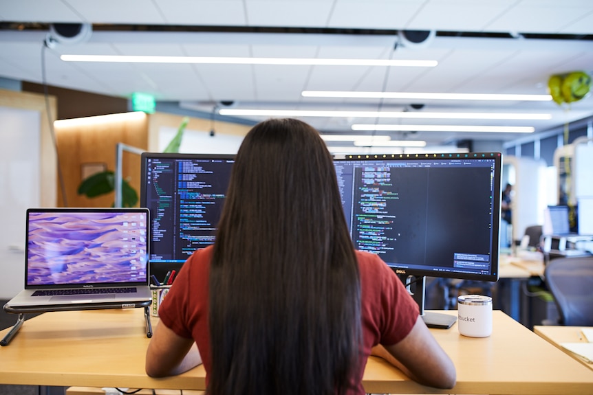 A person with long dark hair works on a computer with software code on two screens.