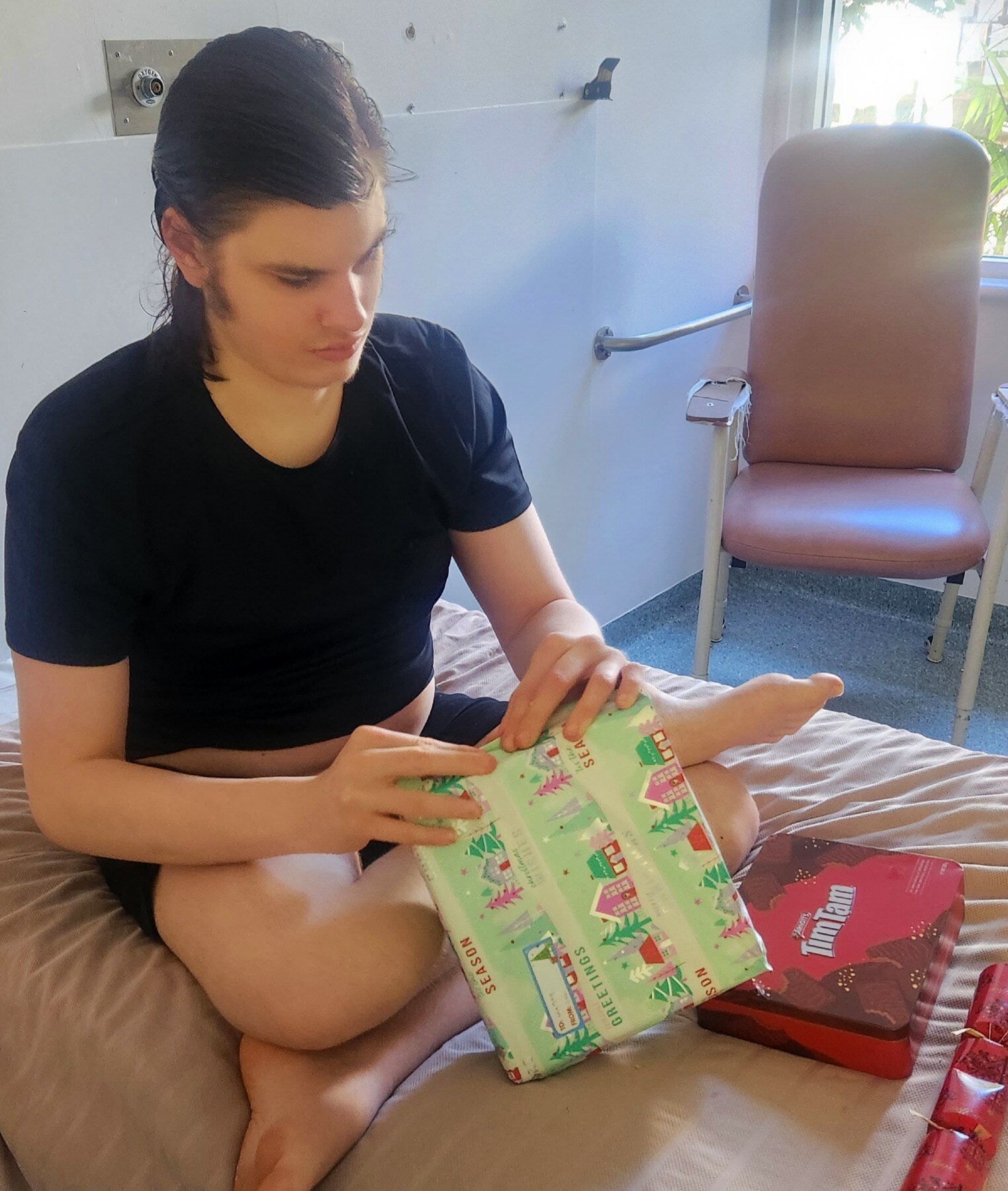 A teenage boy in a black t-shirt sits on a hospital bed opening presents