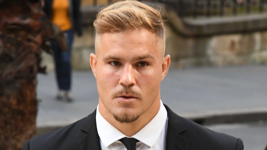 A young man with short blonde hair wears a suit and tie.