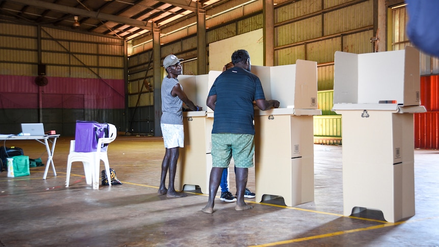 Two Aboriginal men stand at temporary voting booths inside a hall