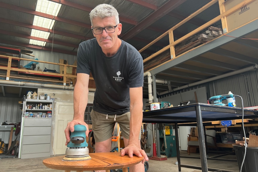 Former AFL player Shaun Smith uses an electric sander while woodworking in a shed.