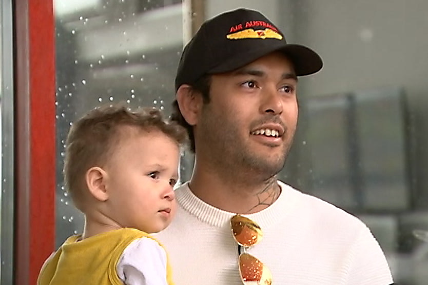 A man in a white jumper, black cap and sunglasses hanging from his neckline talks to reporters while holding a small boy.