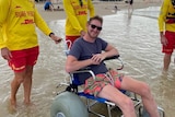 A man in a surf-friendly wheelchair with giant wheels grins as he is wheeled into the water by surf lifeguards.