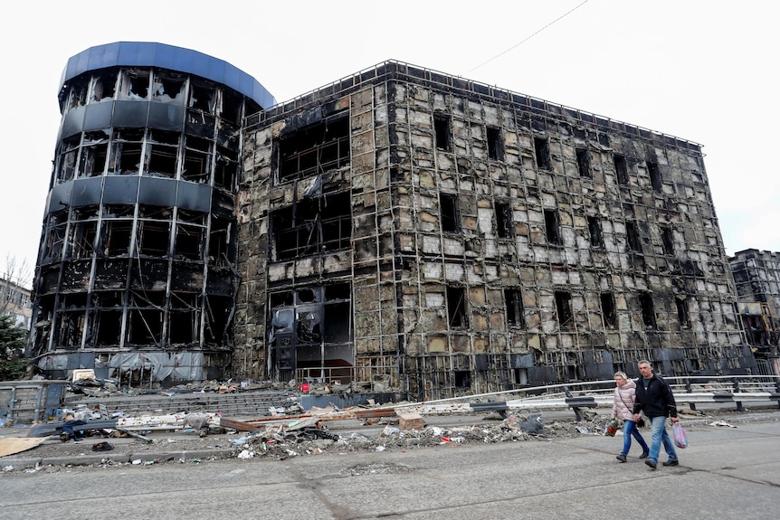 Two people walk past destroyed buildings.